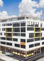 A modern, multi-story commercial building with a large glass facade, yellow accents, and a parking area in front.
