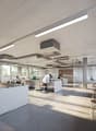 The image shows a modern, open-plan office space with a kitchen area. The space features large windows, allowing natural light to flood in. There are various workstations with desks and chairs, as well as a kitchen counter with what appears to be a dishwa