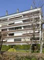 large building with many apartments, multiple floors, white exterior, brown accents, multiple windows, surrounded by greenery