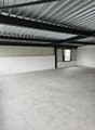 The image shows the interior of a warehouse or industrial space. The ceiling is made of corrugated metal panels, and there are black metal beams supporting the structure. The floor is a plain concrete surface. The space appears to be empty and unfinished,