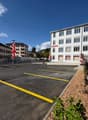 A multi-story commercial building with a large parking lot in front. The building has a modern, white facade with numerous windows. The parking lot is marked with yellow lines and contains several parked cars. There are some decorative plants and flowers