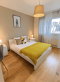 Spacious bedroom with hardwood flooring, white walls, large windows, a double bed with yellow bedding, nightstands, and a pendant light fixture.