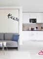 white living room with white walls, gray couch with pillows, white cabinets, wooden microwave, coffee machine, white coffee cups, carpet, white dining table with white chair, pink rug, white table with white cups