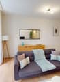Spacious living room with hardwood floors, white walls, large windows, gray sofa, wooden TV stand, and shelving unit. The room also features a balcony or terrace.