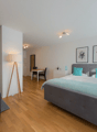 Bedroom with wooden floor, white walls, bed with blue pillows, two nightstands, bedside lamps, wall-mounted pictures, a table and chairs in a corner, a TV, a lamp, and a chair.