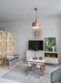 Bright and airy living room with white walls, hardwood floors, and tropical-inspired decor. Features a copper pendant light, a wooden screen divider, a TV stand with storage, and a seating area with a gray sofa and armchair. The room also has potted plant
