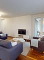 Spacious living room with hardwood floors, white walls, large windows, gray sofa set, coffee table, TV stand, and access to a balcony or terrace