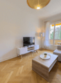 Spacious living room with large windows providing a view, hardwood flooring in a herringbone pattern, white walls, a TV stand, a sofa, and a coffee table with decorative items.