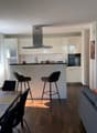 Open kitchen with dining area, living room with couch, dining table, chairs, kitchen island, bar stools, kitchen appliances, window, paintings on wall