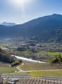 The image shows a picturesque mountain landscape with snow-capped peaks in the background. In the foreground, there are rolling hills covered in vineyards and farmland, with a small town nestled in the valley below. The scene is bathed in warm, golden sun