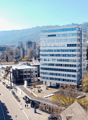 The image shows a cityscape with a mix of modern high-rise buildings and older, smaller structures. There are several multi-story office buildings with glass facades, as well as some residential buildings. The city is surrounded by hills and mountains in