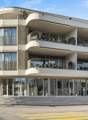 Contemporary multi-storey building with a curved facade, several balconies, glass railings, and a modern glass entrance.