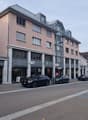 A street with a few parked cars in front of buildings, some with balconies and shops