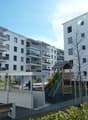 Multi-story apartment building with balconies, parking area, and a children's playground in the foreground.