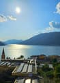 The image shows a scenic view of a lake surrounded by mountains. The lake is reflecting the sunlight, creating a shimmering effect on the water. In the foreground, there is a town with colorful buildings and a church steeple visible. The landscape is lush