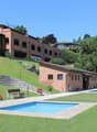 large brick building on the hill, large grassy yard, small pool, small house, benches, patio chairs and tables, large umbrellas