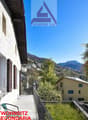 The image shows a two-story residential building with a balcony or terrace, surrounded by palm trees and other greenery. The building has a wooden exterior and large windows. In the background, there are mountains and a blue sky, indicating a scenic locat