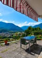 Balcony with a stunning view of the mountains and valley below. The balcony has a striped awning, a table and chairs, and potted plants. The surrounding landscape is lush and green, with mountains in the distance under a blue sky with clouds.