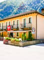 2-story building with a yellow exterior, balconies, and a Swiss flag. The building is surrounded by trees and has a patio area in front with tables and chairs. The building appears to be a hotel or restaurant located in a mountainous, forested area.