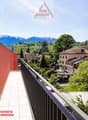 The image shows a balcony or terrace with a view of a small town surrounded by mountains. The buildings in the town appear to be a mix of residential and commercial structures, with trees and greenery visible throughout. The balcony has a red railing and