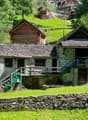 Traditional stone and wooden cabin-style houses in a lush, green mountain setting with a waterfall or stream in the background.