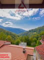 View of a mountainous, forested landscape from a window or balcony. The image shows red-tiled roofs of houses in the foreground, with a backdrop of lush green hills and mountains under a blue sky with clouds.