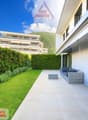 A white building with glass windows, tiled patio leading to a sofa on a terrace, green hedges and grass lawn, and a mountain in the background.