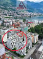 Aerial view of a residential building complex with multiple buildings, surrounded by trees and a lake, mountains in the background.
