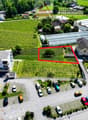 The image shows an aerial view of a residential area surrounded by vineyards and greenery. There is a parking lot visible in the foreground, and several buildings and houses can be seen in the background. The area appears to be a small town or village nes
