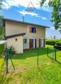 Detached house, beige exterior, green fence, driveway, garden, trees, bushes, clear sky