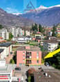 Aerial view of a residential area with a highlighted apartment building, surrounded by other buildings, trees, and mountains in the background.