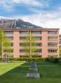 Two story apartment building, white and pink exterior, several balconies, green lawn, bushes, playground, trees, mountains in the background