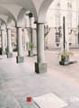 The image shows an outdoor area with arched walkways, columns, and potted plants. The ground is paved with stone tiles, and the surrounding buildings have a historic architectural style. This appears to be a commercial or public space, potentially a plaza