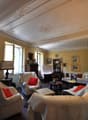 Spacious living room with ornate ceiling, large windows, antique furniture, artwork, and a fireplace