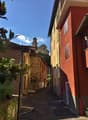 Narrow alleyway with colorful buildings, stone walls, and a church steeple visible in the background. The scene has a charming, historic European feel with lush greenery and a bright, sunny sky.
