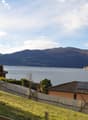 The image shows a scenic view of a lakeside town surrounded by mountains. The town has a mix of residential and commercial buildings with red-tiled roofs. The lake is visible in the foreground, with mountains rising in the background. The landscape appear