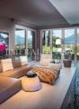 Spacious living room with large windows offering a scenic view of the mountains, modern furniture including a beige sectional sofa, a glass coffee table, and decorative accents such as a pink rabbit sculpture and a wall art piece depicting a cityscape.