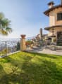 Two-story Mediterranean-style house with a terrace, palm tree, and ornate wrought-iron railing overlooking a grassy yard and mountains in the background. The house has a stone exterior, red tile roof, and decorative architectural details.