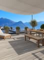 Large wooden deck with outdoor furniture, including lounge chairs and a dining table, with a panoramic view of the mountains and lake in the background. The deck is covered by a canopy and surrounded by potted plants and trees.