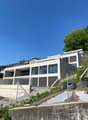 A modern, multi-story building with a balcony or terrace visible, situated on a hillside with lush greenery surrounding it. The building has a sleek, contemporary design with large windows and a gray exterior.