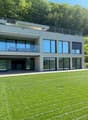 Modern multi-story building with large glass windows, balconies, and a grassy lawn in the foreground.