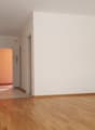 A hallway with wooden floors and white walls, leading to rooms with closed doors and a visible radiator. There's a white sheet on the floor at the end of the hallway.