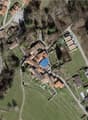 The building is an old medieval-style stone house located in the village. The surrounding area is rural, with houses, farmland, and some trees. A blue arrow points to the main entrance of the building.