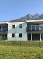 Two-story apartment building with a modern, minimalist design. The building has large windows, balconies, and a garage. It is surrounded by a grassy lawn and has a scenic mountain backdrop.