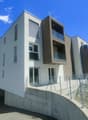 Modern multi-story apartment building with white and dark gray exterior, large windows, and balconies. The building is situated on a raised ground floor with a concrete retaining wall and fencing around the property.