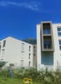 Multi-story apartment building with modern, minimalist design featuring large windows, balconies, and a grassy area with playground equipment in the foreground. The building is set against a backdrop of mountains under a clear blue sky.
