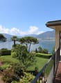 Luxury villa with a panoramic view of a lake surrounded by mountains. The property has a large balcony or terrace overlooking a well-manicured garden with palm trees, shrubs, and colorful flowers. The villa appears to be situated on a hillside, providing