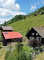 The image shows a picturesque mountain village nestled in a lush, green landscape. The village consists of traditional Swiss-style chalets with wooden facades and slate roofs. There are several buildings with balconies or terraces visible. The village is