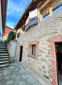 The image shows the exterior of an old stone building with wooden beams and shuttered windows. The building has a stone facade with some brick accents, and there is a set of stairs leading up to the entrance. The surrounding area appears to be a quaint, h