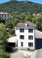 The image shows a small village nestled in a mountainous, forested area. There are several multi-story residential buildings with red tile roofs, surrounded by lush greenery. A road runs through the village, with a parked vehicle visible. The landscape is